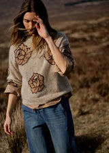 A woman wearing a beige knit sweater with brown floral patterns and blue jeans, standing outdoors in a dry, grassy landscape.