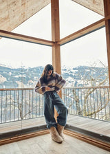 A woman stands on a wooden balcony inside a modern wooden chalet, wearing a fuzzy brown and gray striped sweater, dark wide-leg pants, and beige fur boots, with a snowy mountain landscape visible outside.