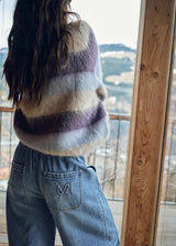 A person viewed from behind wearing a fuzzy striped sweater with horizontal bands of cream, lavender, and dark plum, paired with light blue jeans, standing by a large window with a mountain view.