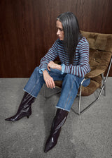 A woman with long dark hair sits in a brown upholstered chair against a dark wood-paneled wall, wearing a blue and red striped long-sleeve top, blue jeans, and dark glossy pointed-toe ankle boots.