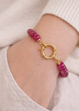 A close-up of a person's wrist wearing a beaded bracelet with faceted magenta-pink beads and a gold clasp, set against a soft cream-colored knit sweater.