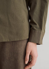 Close-up of a person wearing an olive green long-sleeve shirt with a small embroidered logo on the chest and brown textured trousers, with one hand resting near the hem.