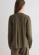 A woman shown from the back wearing an olive green long-sleeve button-up shirt and brown pants against a plain light gray studio background.