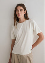 A woman wearing a short-sleeve crewneck t-shirt in off-white and beige trousers, standing against a plain light gray wall.