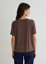 A woman shown from the back wearing a brown short-sleeve crewneck t-shirt and dark blue jeans, standing against a light gray studio background.