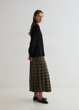 A woman stands in profile wearing a black long-sleeve top and a green and black plaid midi skirt, paired with black loafers, against a plain white studio background.