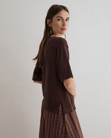 A woman wearing a dark brown short-sleeve top and a brown plaid skirt, photographed from the back against a plain light gray studio background.