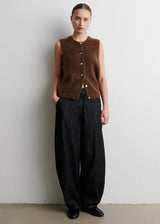 A model stands against a plain light gray wall wearing a brown fuzzy sleeveless button-front vest over a dark charcoal wide-leg pants and black shoes.