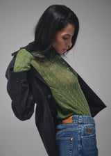 A woman wearing a dark charcoal coat over a green textured knit top and blue jeans, photographed against a plain gray studio background.