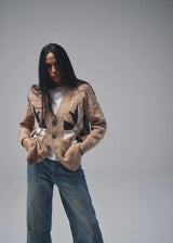 A woman modeling a beige textured jacket with black abstract graphic panels, paired with a white t-shirt and blue jeans, against a solid gray studio background.