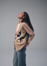 A woman wearing a beige cable-knit cardigan with a black star pattern and a white top, paired with blue jeans, posing against a solid gray studio background.