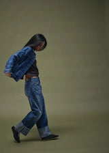 A woman in a studio setting wearing a blue denim jacket, dark gray top, and blue jeans with rolled cuffs, walking against a solid olive-green background.