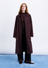 A woman stands against a white studio background wearing a long dark burgundy coat over a dark striped turtleneck, black wide-leg pants, and brown shoes.