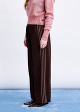 A person wearing dark brown wide-leg trousers with thin vertical cream stripes down the sides, paired with a pink knit sweater and dark loafers, standing against a white wall and blue floor.