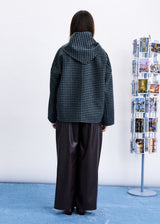 A person standing with their back to the camera, wearing a dark green and black checkered hooded jacket and wide-leg dark brown pants, in front of a white wall with a blue floor. A clothing display stand is visible on the right.