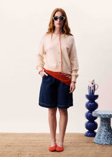 Woman wearing a beige cardigan, red shirt, blue shorts, and sunglasses indoors.