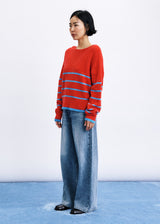 A model stands in a studio wearing a red knit sweater with horizontal blue stripes and wide-leg blue jeans against a white backdrop with a blue floor.