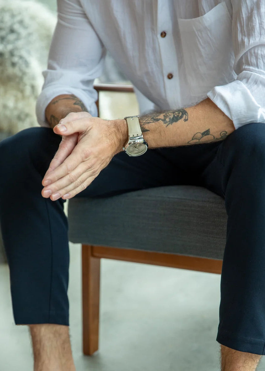 Person sitting on a chair wearing a white shirt and dark pants, with tattoos visible on arms.