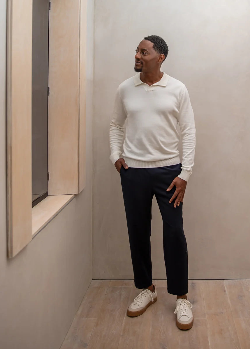 Man wearing a white sweater and dark pants standing in a minimalistic room.