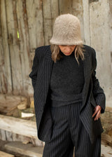 A person wearing a dark pinstripe blazer and matching trousers with a dark gray knit top, a beige fuzzy bucket hat, and looking down, standing in front of a rustic wooden wall.