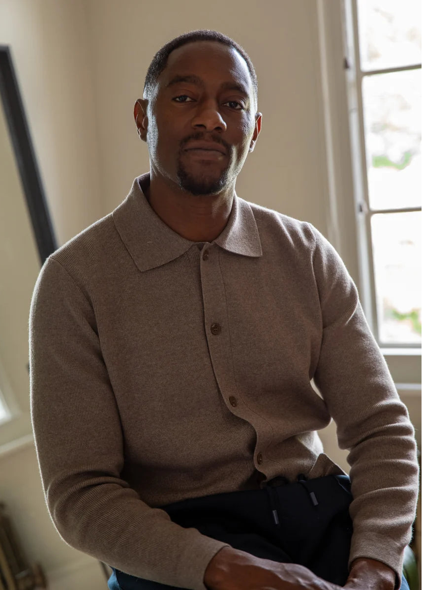 A man wearing a beige long-sleeve knit polo cardigan sweater and dark trousers, seated indoors near a window.