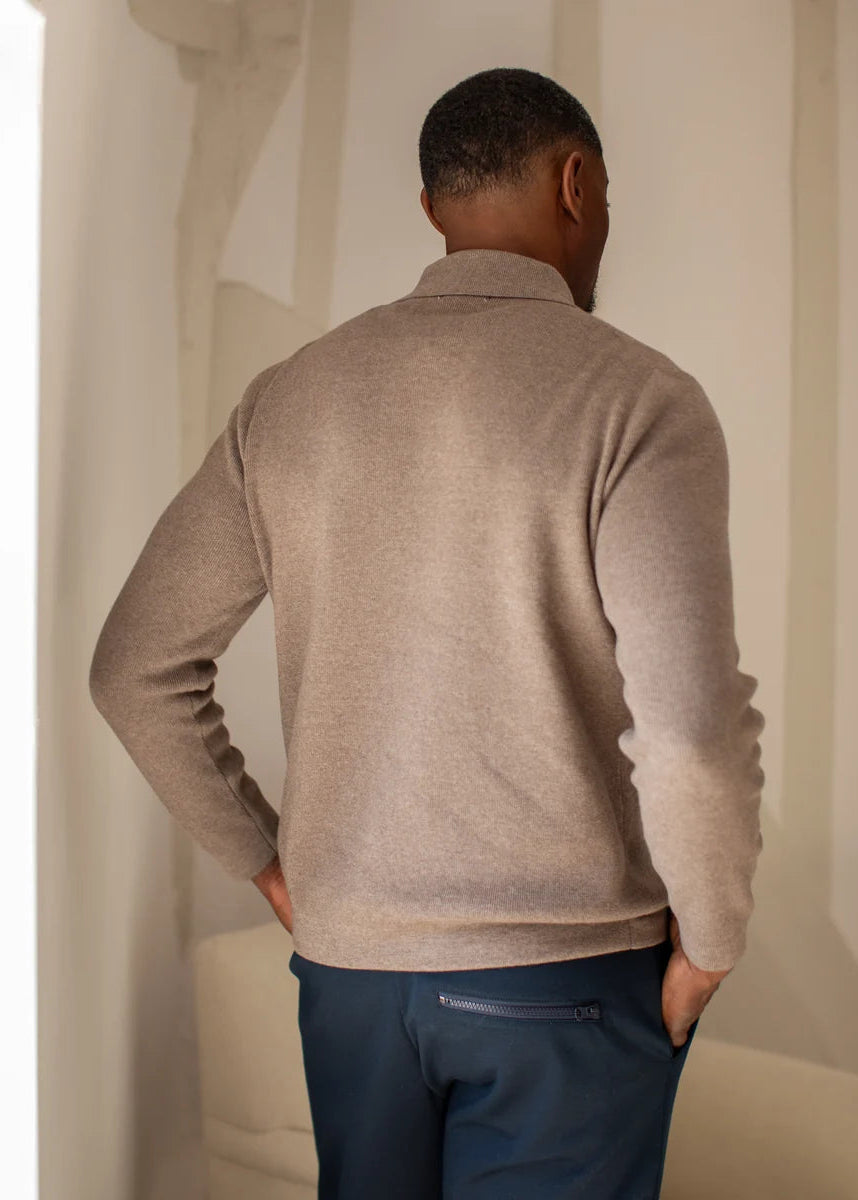A man viewed from behind wearing a light taupe knit quarter-zip sweater and navy trousers, standing in a softly lit indoor setting.