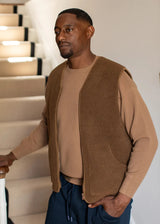A man wearing a brown knit sleeveless vest over a tan crewneck sweater and navy drawstring trousers, standing indoors near a staircase.