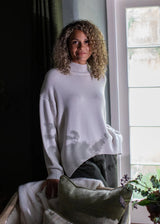 A woman with curly blonde hair wearing a white knit mock-neck sweater and dark gray pants, leaning against a window in a dimly lit room with a pillow in the foreground.