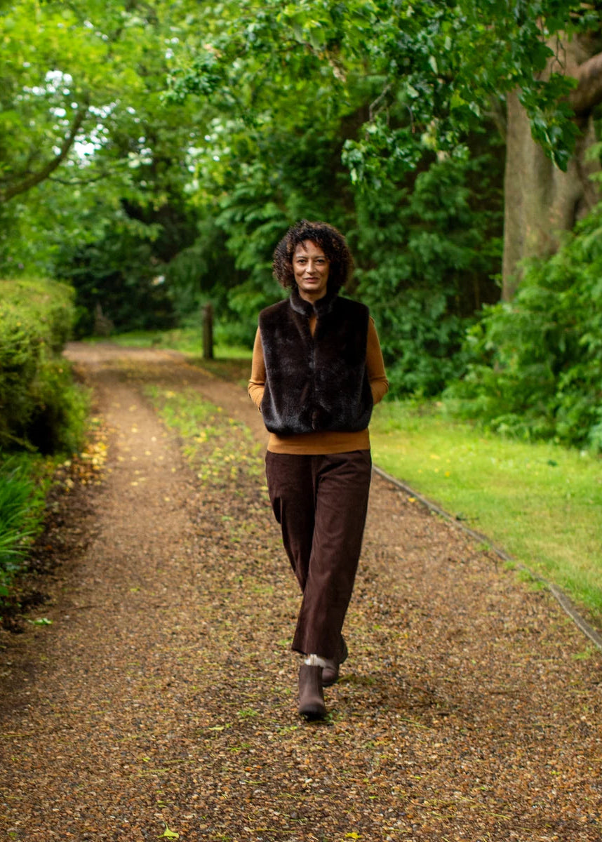A woman walking on a leaf-strewn path in a lush green park, wearing a dark brown faux-fur vest over a tan top and dark brown wide-leg trousers.