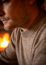A close-up side profile of a man wearing a warm taupe knit sweater, with a glowing fireplace blurred in the background.