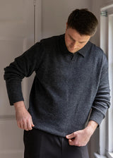 A man wearing a dark charcoal gray knit polo sweater and dark trousers, standing in a room with a white paneled door and a window.