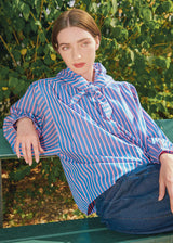A woman wearing a blue and white striped blouse with a ruffled tie-neck collar, sitting on a green park bench with leafy bushes in the background.