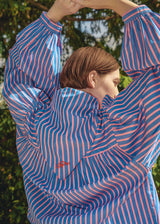 A model wearing a blue and pink vertically striped oversized shirt, photographed outdoors with their arms raised above their head.