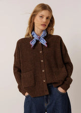 A woman wearing a brown ribbed button-up cardigan with front pockets, styled with a blue and red patterned neck scarf and dark blue jeans, against a plain light background.