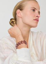 A woman in a white blouse wearing a chunky, chunky purple-brown chain-link bracelet, shown in a close-up profile against a light gray background.
