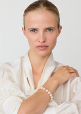 A woman with her hair pulled back wears a white satin blouse and a light pink beaded bracelet, photographed against a clean white studio background.
