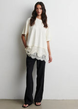 Woman wearing a white top with lace trim and black pants against a plain background