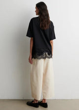 Woman wearing a black top with lace detail and white pants against a plain background