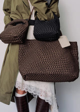 A person wearing an olive green coat and white skirt carries three woven bags: a large brown tote bag, a smaller brown shoulder bag, and a black woven bag, against a plain light gray background.