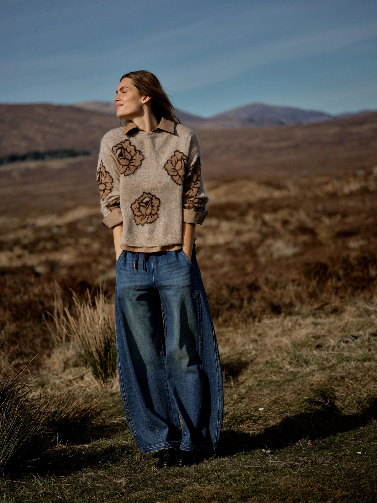 Woman standing in a field with mountains in the background wearing a patterned sweater and jeans.