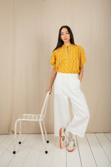 Woman in a yellow patterned top and white pants standing next to a small chair against a beige wall.
