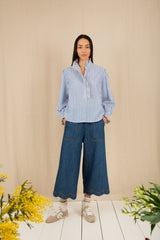 Woman wearing a blue and white striped shirt and blue jeans standing against a beige wall with yellow flowers on a wooden floor.