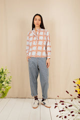 Woman wearing a patterned shirt and jeans standing against a beige wall with decorative plants.