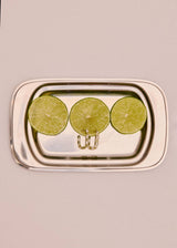 A rectangular silver metal tray with rounded corners and a small hanging loop at the bottom center, holding three sliced lime wedges on a plain light beige background.