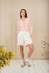 Woman wearing a pink checkered shirt and white shorts standing against a beige wall with floral decorations.