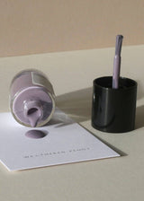 A bottle of lavender nail polish lies on its side with polish dripping onto a white card, next to a black cup holding the brush. The card reads 'WEATHERED PENNY'.
