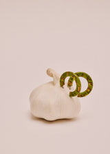 A pair of green and gold patterned hoop earrings displayed on a white garlic bulb against a solid pale pink background.
