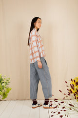 Woman wearing a patterned shirt and jeans standing against a beige wall with plants and flowers on a light wooden floor.