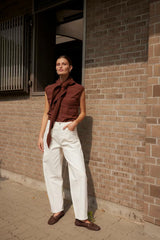 Woman standing against a brick wall wearing a brown top and white pants.
