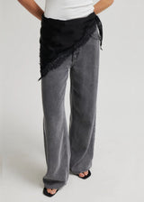 A person wearing wide-leg gray jeans with a black asymmetrical wrap-style top or sash featuring lace trim, paired with a white top and black sandals, photographed against a light gray studio background.
